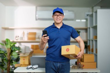 Bir teslimat işçisi lojistik ofisinde elinde paketler ve akıllı bir telefonla müşterilere yardım etmeye hazır bekliyor. Modern dağıtım hizmetlerini sunar.