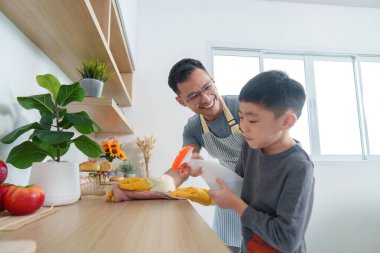 Baba, oğluna mutfak yüzeylerini temizlemede rehberlik ediyor, takım çalışmasını ve evde bakım sorumluluğunu teşvik ediyor..
