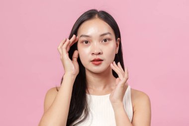 Işıl ışıl gülümseyen genç bir kadın yüzüne hafifçe dokunuyor, pembe bir arka plana karşı güzelliği ve özgüveni vurguluyor..