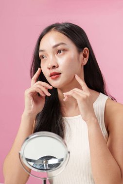 Genç bir kadın cildini incelikle inceler cilt ürünlerini uygularken, güzelliği ve özgüveni vurgulayarak pembe bir zemine karşı.