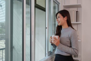 Elinde kahve fincanı tutan ve pencereden dışarı bakan bir kadın, sakin bir sabah rutininde gününü yansıtıyor..