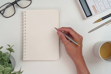 Beyaz bir masanın üzerinde defter, kalem ve kahve fincanı bulunan temiz ve düzenli bir çalışma alanı..