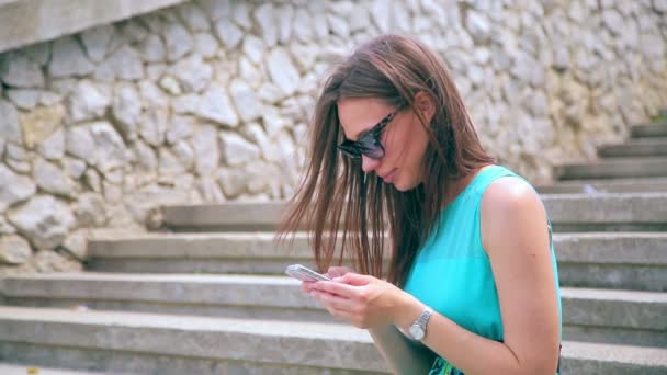 Atrakcyjna dziewczyna wpisując tekst na telefon siedzi na starych schodach. Zbliżenie — Wideo stockowe