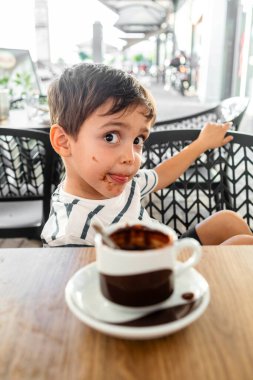 Neşeli küçük çocuk, yüzü çikolata lekeleriyle kaplı bir fincan sıcak çikolatanın tadını çıkarıyor..