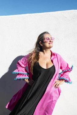 Güneş ışığında poz veren zarif genç bir kadın pembe kalp şeklinde güneş gözlüğü, siyah elbise ve pembe fırfırlı bir bluz giyerek beyaz bir duvarın önünde duruyor..