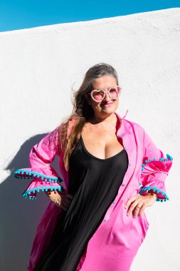 Güneş ışığında poz veren zarif genç bir kadın pembe kalp şeklinde güneş gözlüğü, siyah elbise ve pembe fırfırlı bir bluz giyerek beyaz bir duvarın önünde duruyor..