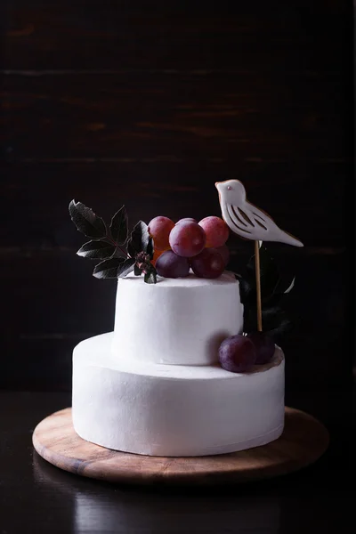 Biały tort dwu warstwowych z winogron ptak i pozostawia na ciemnym tle drewniane z ciemności światło — Zdjęcie stockowe