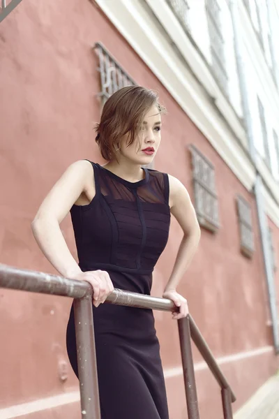 Modello Vogue nel vestito nero esterno vicino edificio vicino alla recinzione — Foto Stock