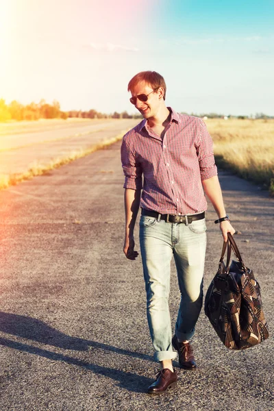 Stylish man in the jeans and sunglasses with the bag walking on the road outdoor
