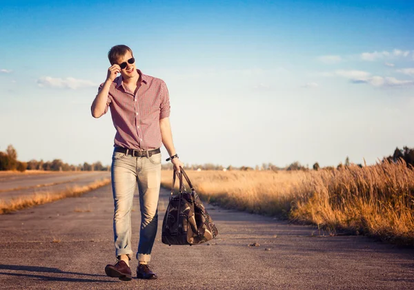 Hombre con estilo en los pantalones vaqueros y gafas de sol con la bolsa caminando en el camino al aire libre — Foto de Stock