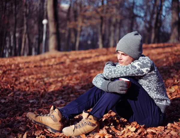 Bello elegante felice giovane nel maglione e cappello con la tazza di tè o caffè nel parco autunnale — Foto Stock