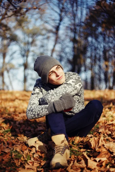 Bonito elegante jovem feliz no suéter e chapéu com a xícara de chá ou café no parque de outono — Fotografia de Stock
