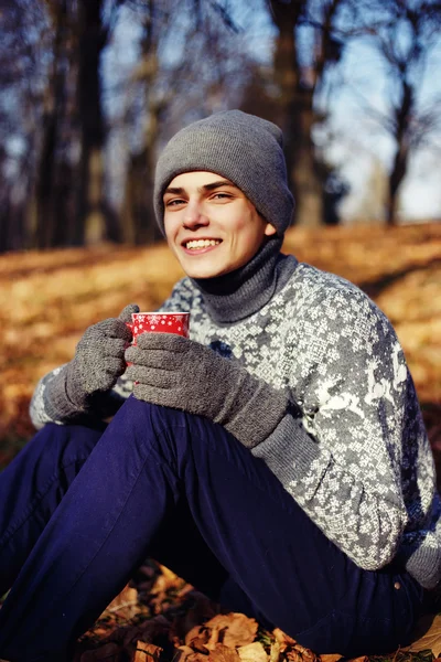 Гарний стильний щасливий молодий чоловік в светрі і капелюсі з чашкою чаю або кави в осінньому парку — стокове фото
