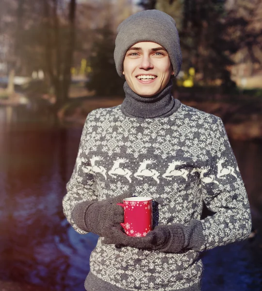 Гарний стильний щасливий молодий чоловік в светрі і капелюсі з чашкою чаю або кави в осінньому парку біля води — стокове фото