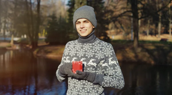 Гарний стильний щасливий молодий чоловік в светрі і капелюсі з чашкою чаю або кави в осінньому парку біля води — стокове фото