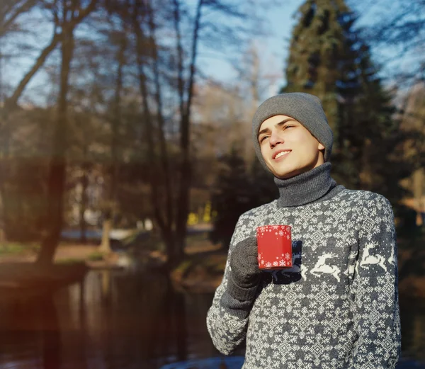 Szép stílusos boldog fiatal férfi pulóver és a csésze tea vagy kávé a őszi Park közelében víz kalap — Stock Fotó