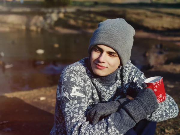Κομψό ευτυχισμένη Oμορφος νέος στο πουλόβερ και καπέλο με το φλιτζάνι τσάι ή καφέ στο φθινοπωρινό πάρκο κοντά στο νερό — Φωτογραφία Αρχείου