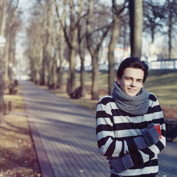 Szép stílusos boldog fiatalember a pulóver és a sálat a csésze tea vagy kávé a őszi parkban — Stock Fotó