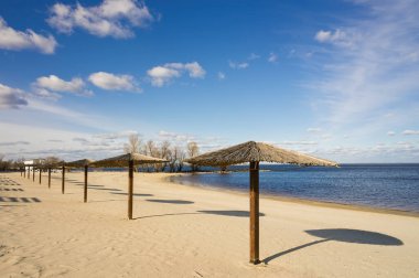 Ukrayna, Cherkasy yakınlarındaki Dinyeper nehri kıyısındaki kumsalda birkaç boş saman şemsiyesi beyaz bulutlu açık mavi gökyüzü altında gölgeler bırakarak sükunet ve seyahat temalarını çağrıştırıyor.