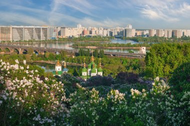 Vydubychi Manastırı ve Dinyeper Nehri Ukrayna 'nın başkenti Kyiv' den akıyor. Etrafında çiçek açan leylak çalıları ve berrak mavi gökyüzünün altında modern bir şehir manzarası var.