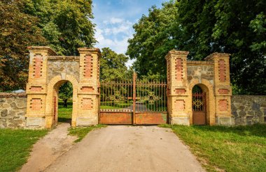 Süslü demir kapı, Samchyky, Khmelnytskyi bölgesinde yemyeşil ağaçlarla çevrili tarihi bir sarayın girişini gösteren tuğla ve taş sütunlarla süslenmiş.