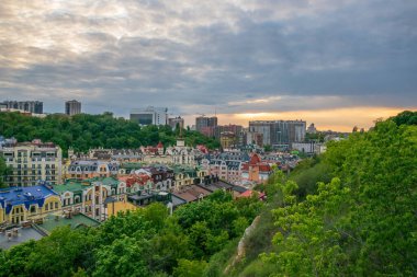 Vozdvyzhenka bölgesinin renkli evlerini gösteren Kyiv şehir manzarası modern binalar ve gün batımında bulutlu bir gökyüzünün altında parlak bir ufuk ile yemyeşil ağaçların arasına kurulmuştur.