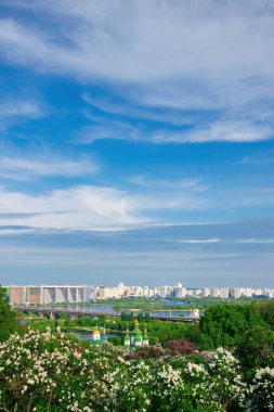 Yeşil kubbeleri ve altın kubbeleri ile Vydubychi Manastırı Dinyeper nehrine ve Ukrayna 'nın modern ufuk çizgisine bakan açık mavi gökyüzünün altında çiçek açan leylak çalılarının arasında duruyor.