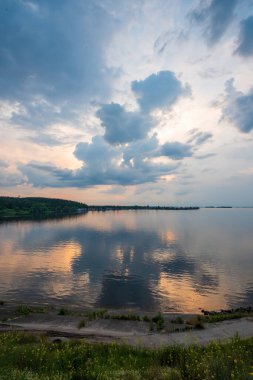 Dinyeper nehri, uzak yeşil bir orman kıyı şeridi ve sakin bir su yüzeyi ile akşam gökyüzünün renkli bulutlarını yansıtıyor ve Ukrayna 'da sakin bir manzara oluşturuyor.