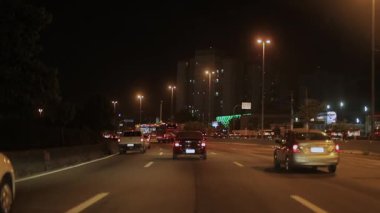 Şehir otobanında gece trafiği, sokak lambaları altında arabalar, arka planda şehir silüeti, hafif araç akışı ve uzak yüksek binalarla aydınlatılmış yol.