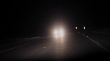 Yoğun farları olan araba, uzak bir gece yolunda, farklı yönlerden yaklaşıyor. Işıklar yanıp sönüyor ve kırsal alanda izole sürüş koşulları var.