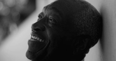 Afro-Amerikalı yaşlı adam yukarıya bakarken sıcak bir şekilde gülümsüyor, siyah beyaz bir duvara yaslanıyor, memnuniyet, dinginlik ve pozitiflik yayıyor.