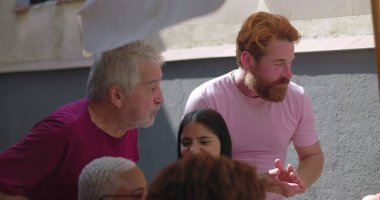 Çeşitli arkadaşlar canlı bir yemek sırasında dışarıda alkışlıyor ve gülüyor, bir şemsiye altında yapılan çok kültürlü bir toplantıda bağlantıyı, kutlamayı ve eğlenceyi vurguluyorlar.