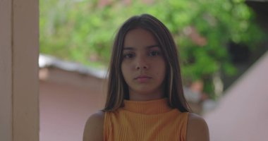 Tarafsız bir ifadeyle dışarıda duran genç kız, doğrudan kameraya bakıyor, sakin ve düşünceli, canlı yeşil arka plan, barışçıl ve içsel an