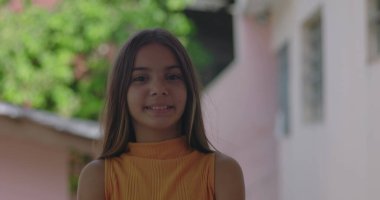 Genç kız kameraya sıcak bir şekilde gülümsüyor, arka planda yeşillik, gerçek ve mutlu bir ifade, doğal ve rahat bir ortamda neşeli bir bağlantı.