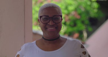 Gözlüklü gülümseyen kadın doğrudan kameraya bakıyor, doğal ortamında, neşeli ve kendine güvenen ifade, bireyselliği kucaklayan ve enerjik kişiliğe sahip.