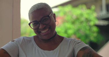 Afrikalı Latin kökenli bir kadın neşeyle dışarıda gülüyor, kafası arkaya eğik, kısa beyaz saçlı ve gözlüklü, arka planda canlı yeşillik, saf mutluluk anı.