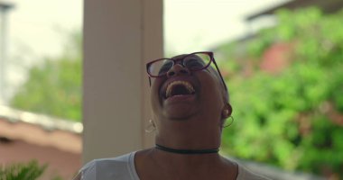 Afrikalı Latin kökenli bir kadın neşeli bir şekilde gülüyor, elleri anlamlı bir anda kaldırılmış, kısa beyaz saçlı ve gözlüklü, açık havada, canlı doğal bir geçmişi var.