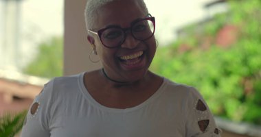 Afrikalı Latin kökenli bir kadın neşeli bir şekilde gülüyor, elleri anlamlı bir anda kaldırılmış, kısa beyaz saçlı ve gözlüklü, açık havada, canlı doğal bir geçmişi var.