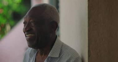 Afrika kökenli gülümseyen yaşlı bir adam, yan tarafa bakıyor, açık havada, canlı bir ortamda, sıcak, mutluluk saçan, kendine güvenen, yaklaşılabilir bir tavırda.