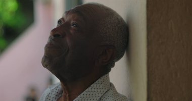 Afrika kökenli yaşlı bir adam duvara yaslanmış, sakin ve düşünceli bir ifadeyle yukarıya bakıyor, arka planda canlı yeşillikle, sakin ve yansıtıcı.