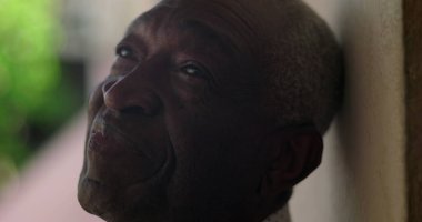 Afrika kökenli yaşlı bir adam duvara yaslanmış, sakin ve sakin bir ifadeyle yukarıya bakıyor, açık hava, canlı arka plan, yansıtıcı ve barışçıl