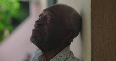 Afrika kökenli yaşlı bir adam duvara yaslanmış, sakin ve düşünceli bir ifadeyle yukarıya bakıyor, arka planda canlı yeşillikle, sakin ve yansıtıcı.