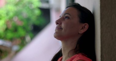 Bir kadın duvara yaslanıyor, sakin bir ifadeyle yukarı bakıyor, bulanık yeşil bir arka plana yakın çekim, yansıma ve huzur anı