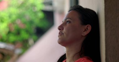 Duvara yaslanmış bir kadın, alnına hafifçe yaslanmış bir el, pişmanlık ve duygusal ifade, bulanık yeşil arka plana yakın çekim, açık havada yansıma anı