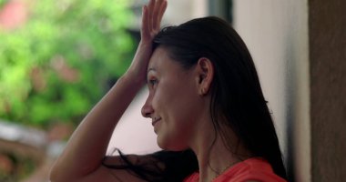 Duvara yaslanmış bir kadın, alnına hafifçe yaslanmış bir el, pişmanlık ve duygusal ifade, bulanık yeşil arka plana yakın çekim, açık havada yansıma anı