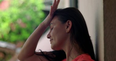 Duvara yaslanmış bir kadın, alnına hafifçe yaslanmış bir el, pişmanlık ve duygusal ifade, bulanık yeşil arka plana yakın çekim, açık havada yansıma anı