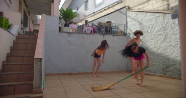 Avluda süpürgeyle oynayan çocuklar, kardeş gülüşü, kaygısız eğlence, zıplama, aile bağları, neşeli enerji, neşeli an, açık hava heyecanı. Hayatta basit şeyler.