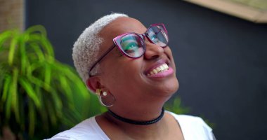 Afro-Amerikalı bir kadın, parlak bir gülümsemeyle, düşünceli ve neşeli bir ifadeyle, yeşillik ve kendine güvenle dışarı bakıyor.