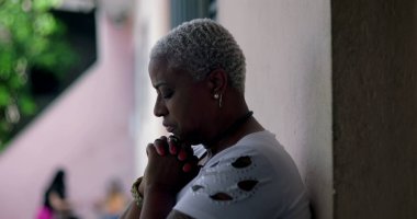 Derin düşüncelere sahip bir kadın, duvara yaslanmış, elleri birbirine kenetlenmiş dua, duygusal rahatlama, Afrika kökenli, yansıtıcı an, açık hava