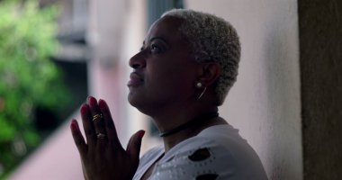 Umut dolu bir kadın, duvara yaslanmış, elleri dua ederken kenetlenmiş, Afrika kökenli, açık hava manzaralı, iyimser bir yüz ifadesi olan.